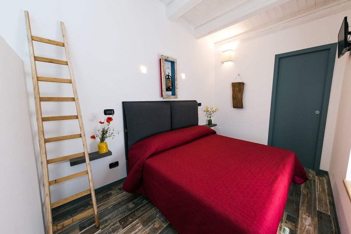 Un dormitorio con cama, una escalera, una estantería con flores, un cuadro en la pared y una puerta.