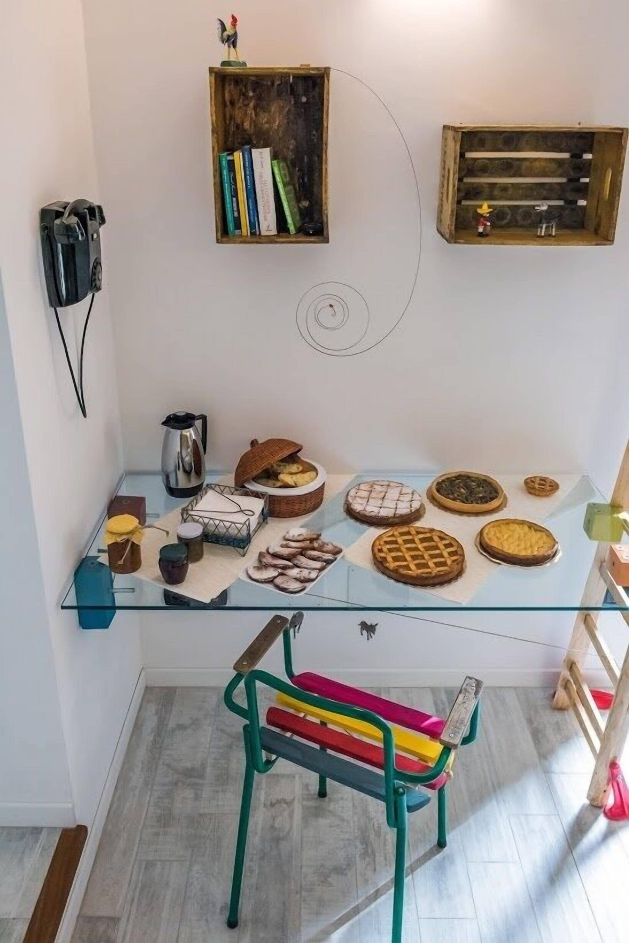 Um quarto com uma mesa de vidro exibindo diversos doces, uma cadeira colorida e estantes de madeira com livros e uma pequena estatueta.
