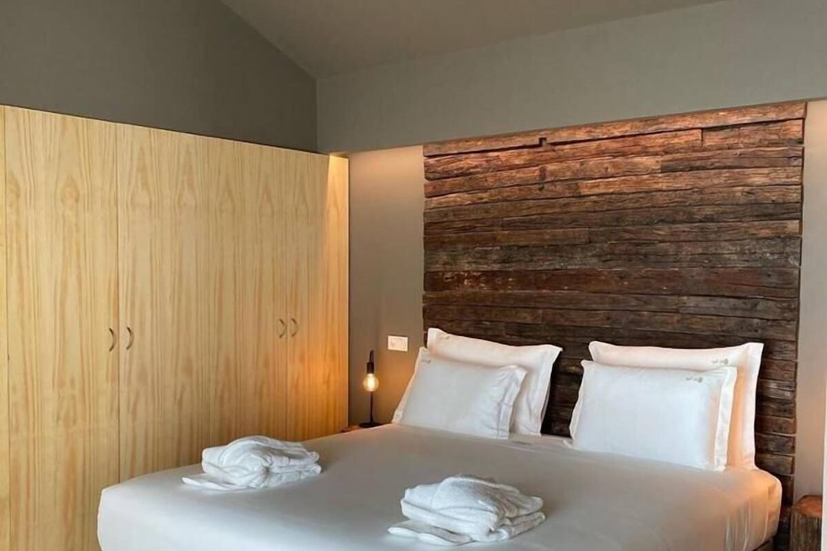 Une chambre moderne avec une armoire en bois, un grand lit avec du linge de lit blanc et une tête de lit à l’aspect pierre.