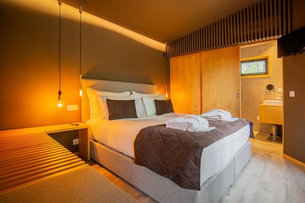 A modern hotel room with a large bed, a wooden headboard, a bedside table, a nightstand, a built-in bench, a small window, a mirror, and a bathroom with a sink and toilet.