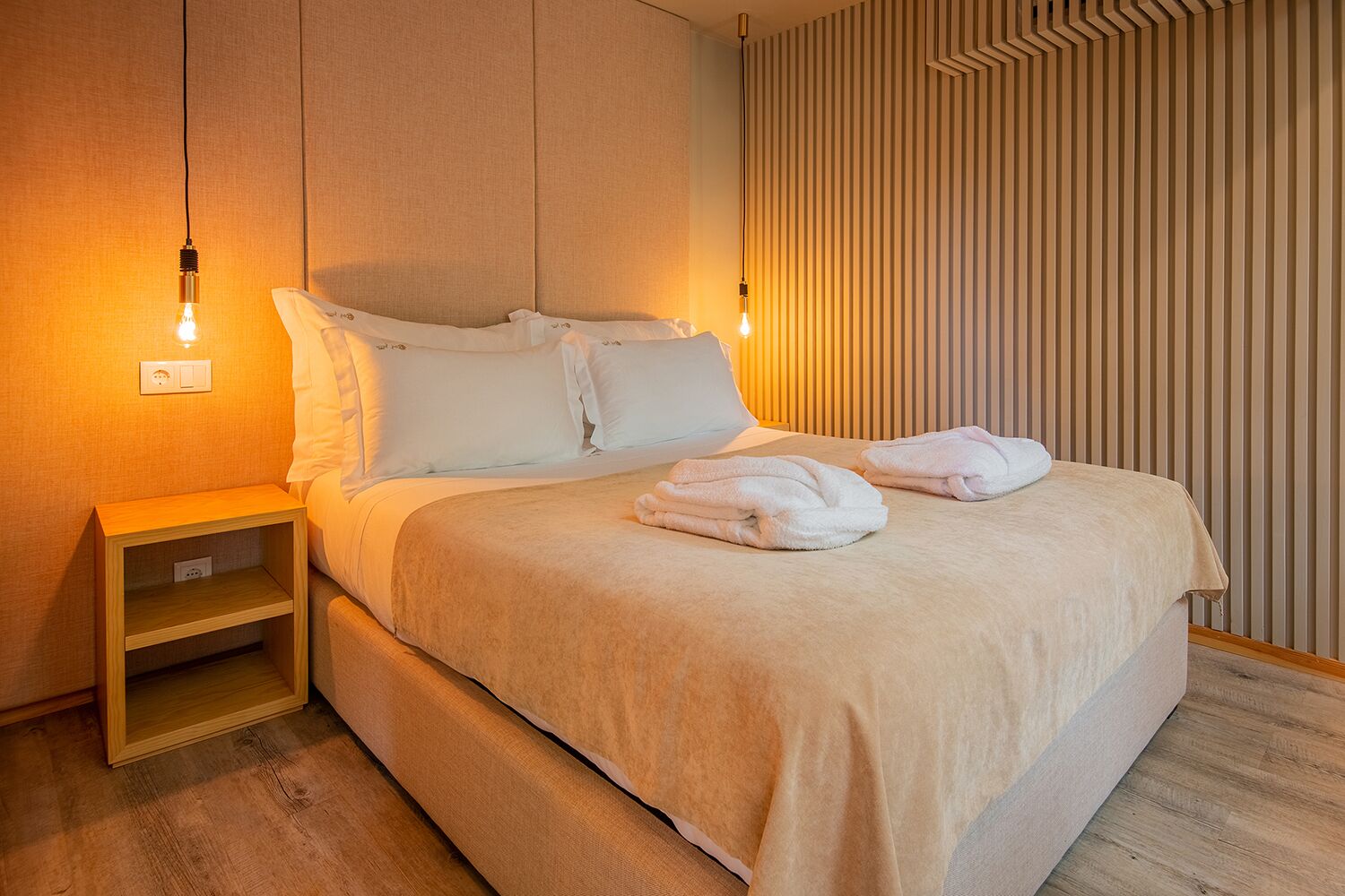 Una cama bien hecha con sábanas blancas y dos toallas dobladas, una mesita de noche de madera con un enchufe, y una pared con listones de madera verticales.