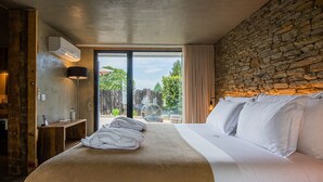 Quarto com uma cama grande, parede de pedra, um banco de madeira, um abajur e vista para o verde lá fora.