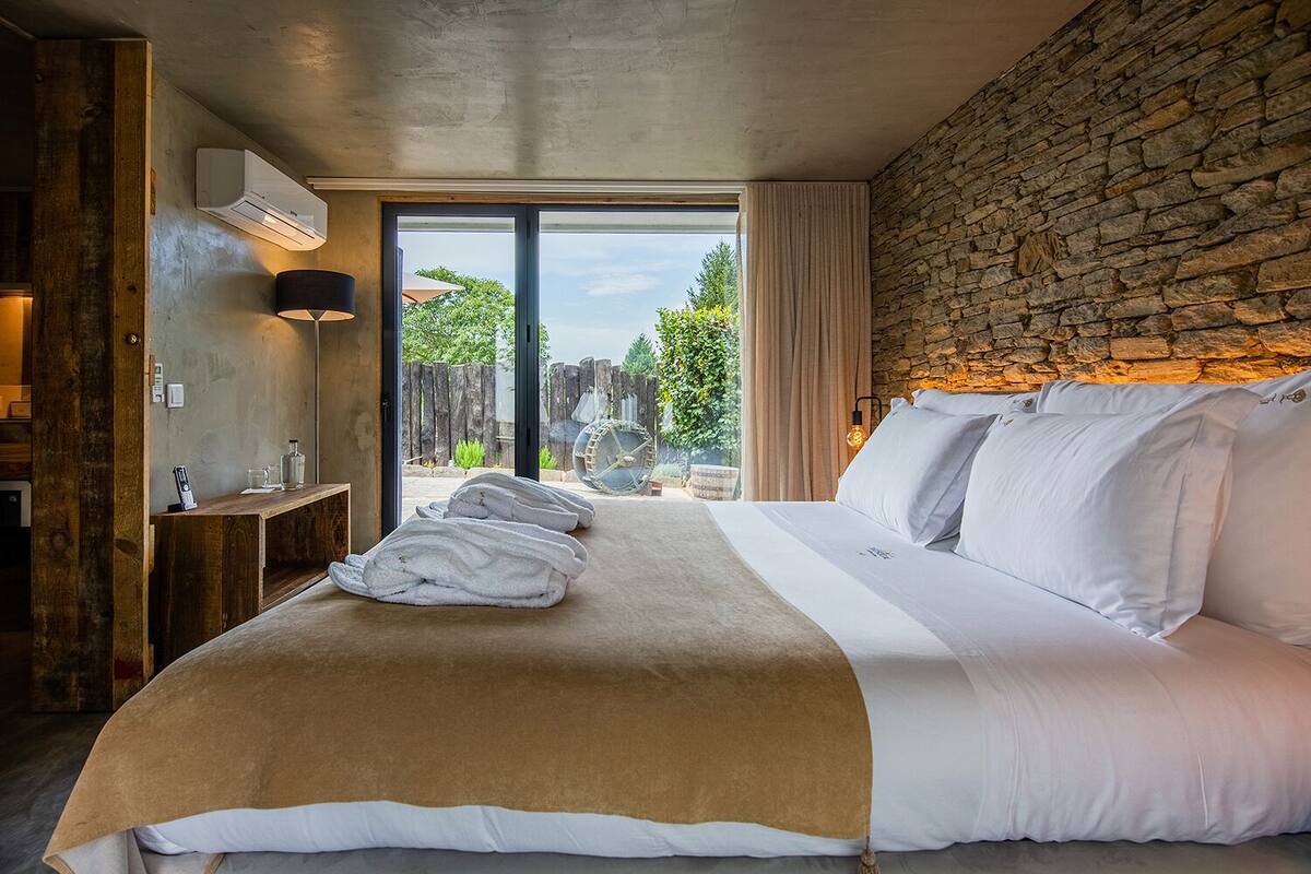 Une chambre avec un grand lit, un mur en pierre, un banc en bois, une lampe et une vue sur la verdure à l’extérieur.