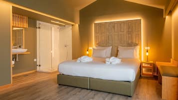 A bedroom with a large bed, a wooden headboard, bedside tables with lamps, a wooden bench, and a bathroom with a glass shower enclosure.