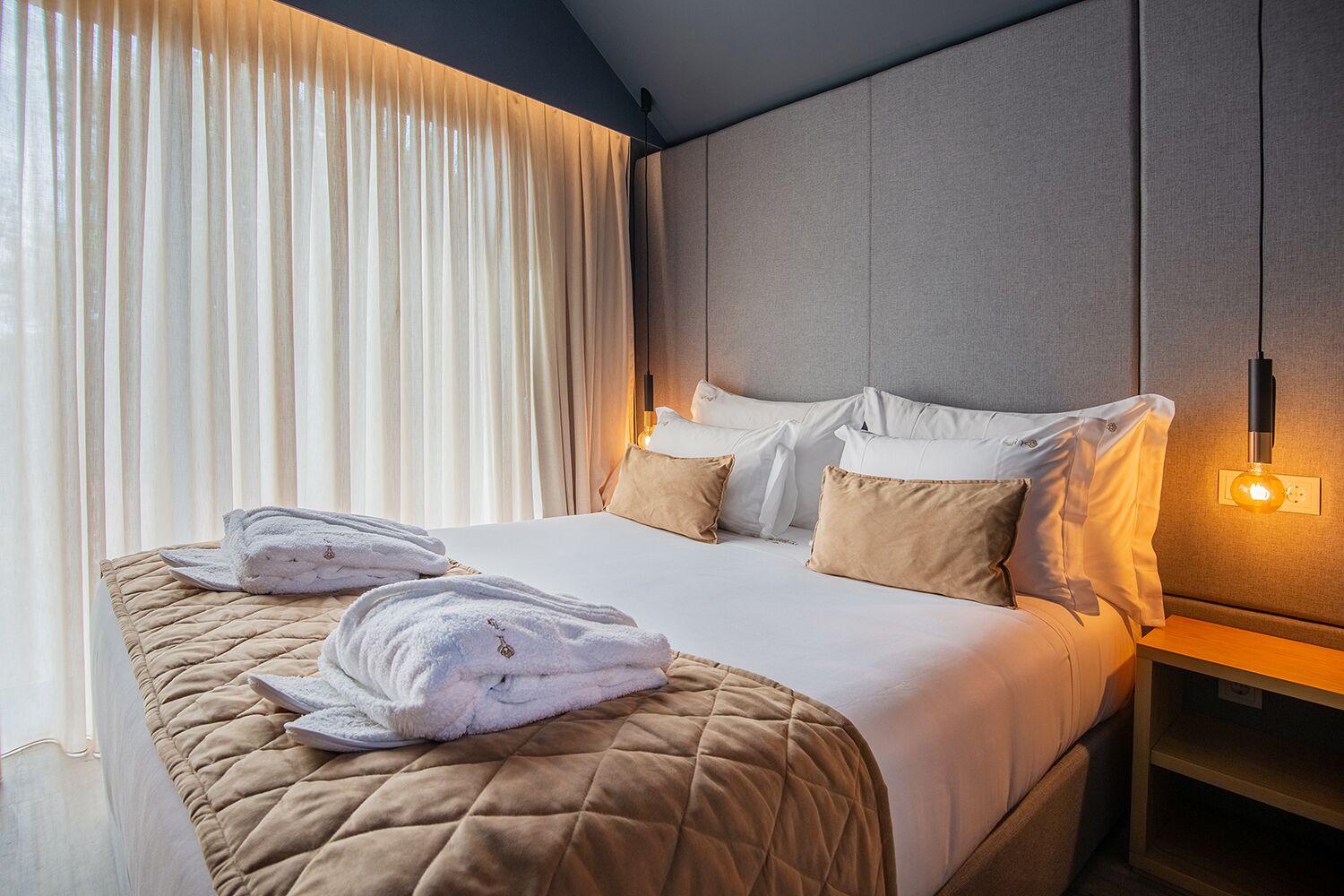 Quarto de hotel com cama, duas almofadas, mesa de cabeceira, abajur e janela com cortinas.