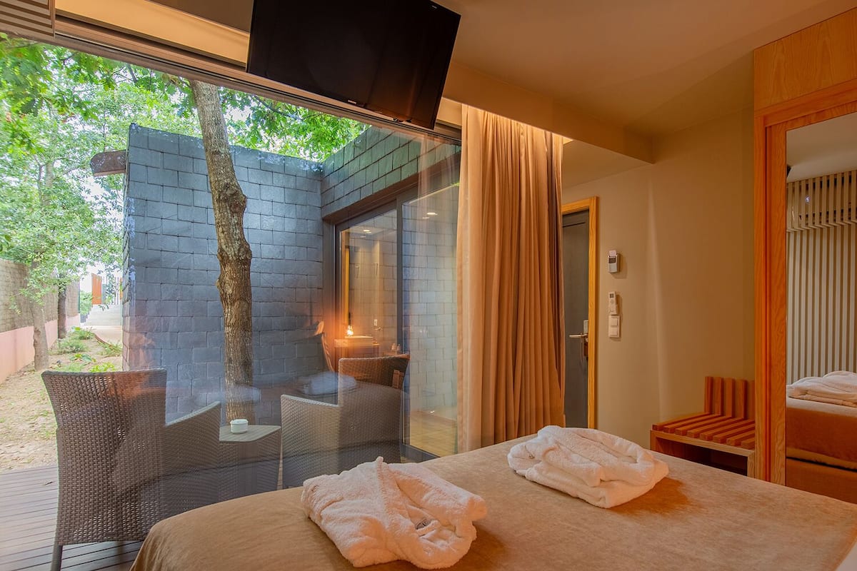 Une chambre d’hôtel comprenant un lit, une télévision, un mur en verre, une fenêtre, une porte et un petit coin salon avec une chaise et une table.