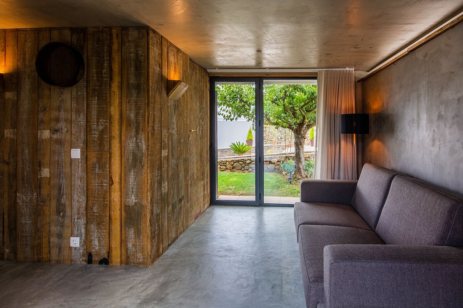 A modern interior with a wooden accent wall, a grey sofa, and a large sliding glass door leading to an outdoor garden.
