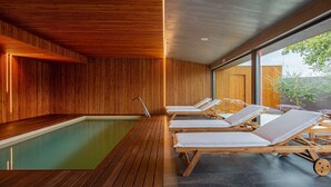 Área interna de piscina moderna, com ripas de madeira nas paredes, piscina e cadeiras de descanso.
