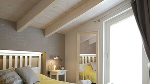 A bedroom with a wooden ceiling, a bed with yellow bedding, a bedside table with a lamp, a mirror, and a window with curtains. - Vicolo Fiore Affittacamere (Matera)
