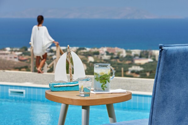 A poolside table with a pitcher of water and a glass, a chair, and a view of the sea and buildings in the background.
