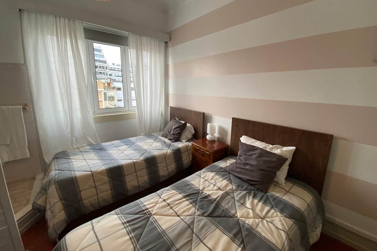 Ein Hotelzimmer mit zwei Betten, rautenmusterigen Wänden, einem Fenster mit Vorhängen und Blick auf Gebäude.