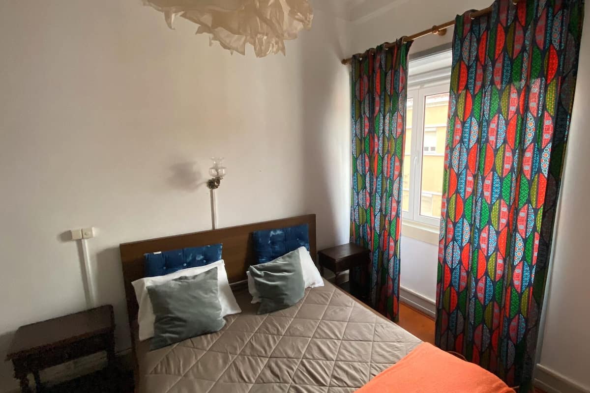 Ein Schlafzimmer mit Bett, Kissen, Nachttisch, Lampe und einem Fenster mit Vorhängen.