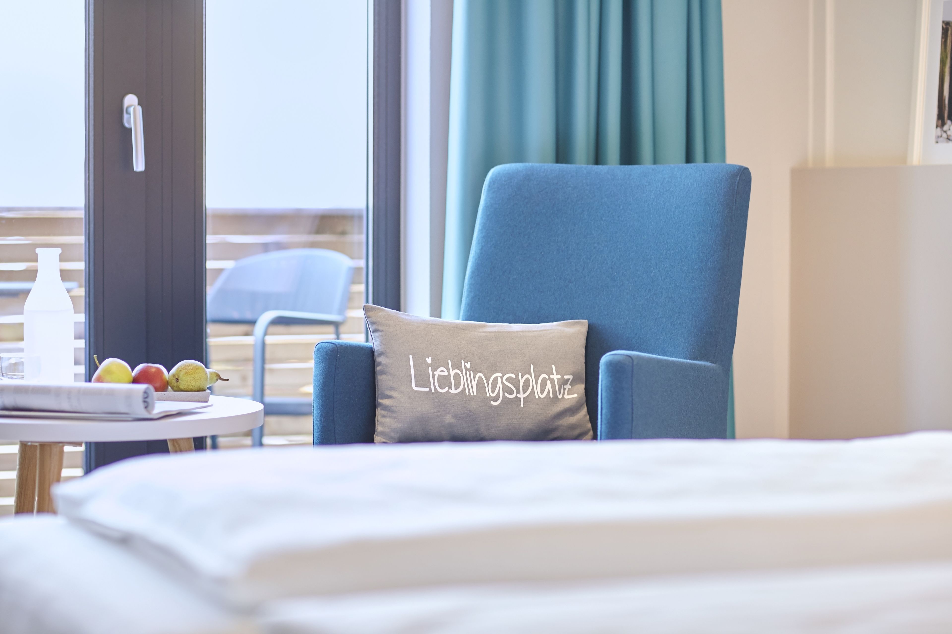 Une chambre d’hôtel avec un lit, un fauteuil bleu, une table avec des fruits et une fenêtre avec des rideaux.