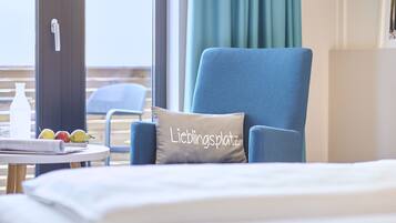 A hotel room with a bed, a blue armchair, a table with fruits, and a window with curtains.