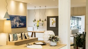 A reception area with a wooden desk, a large painting, pendant lights, and a view into the living area. - Strandkind - Dein Ostseehotel (Neustadt in Holstein)
