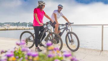 Dos personas en bicicleta junto a la orilla del agua, con una barandilla y flores al frente.