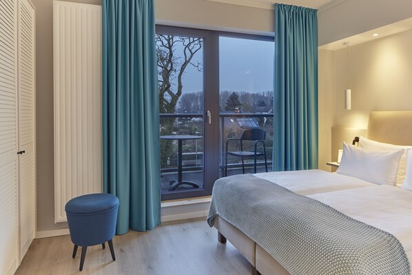 Habitación de hotel con cama, un taburete pequeño, una ventana con cortinas y vistas al exterior.