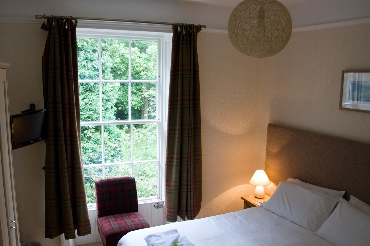 A hotel room with a bed, a chair, a window with curtains, a lamp, and a wall-mounted TV.