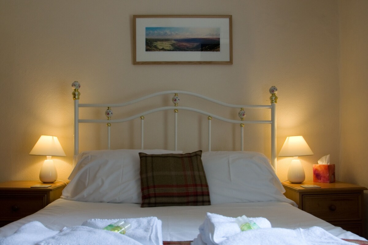 A neatly made bed with white linens, two pillows, and a plaid throw. There are two bedside lamps on wooden nightstands, and a framed picture hangs above the bed.