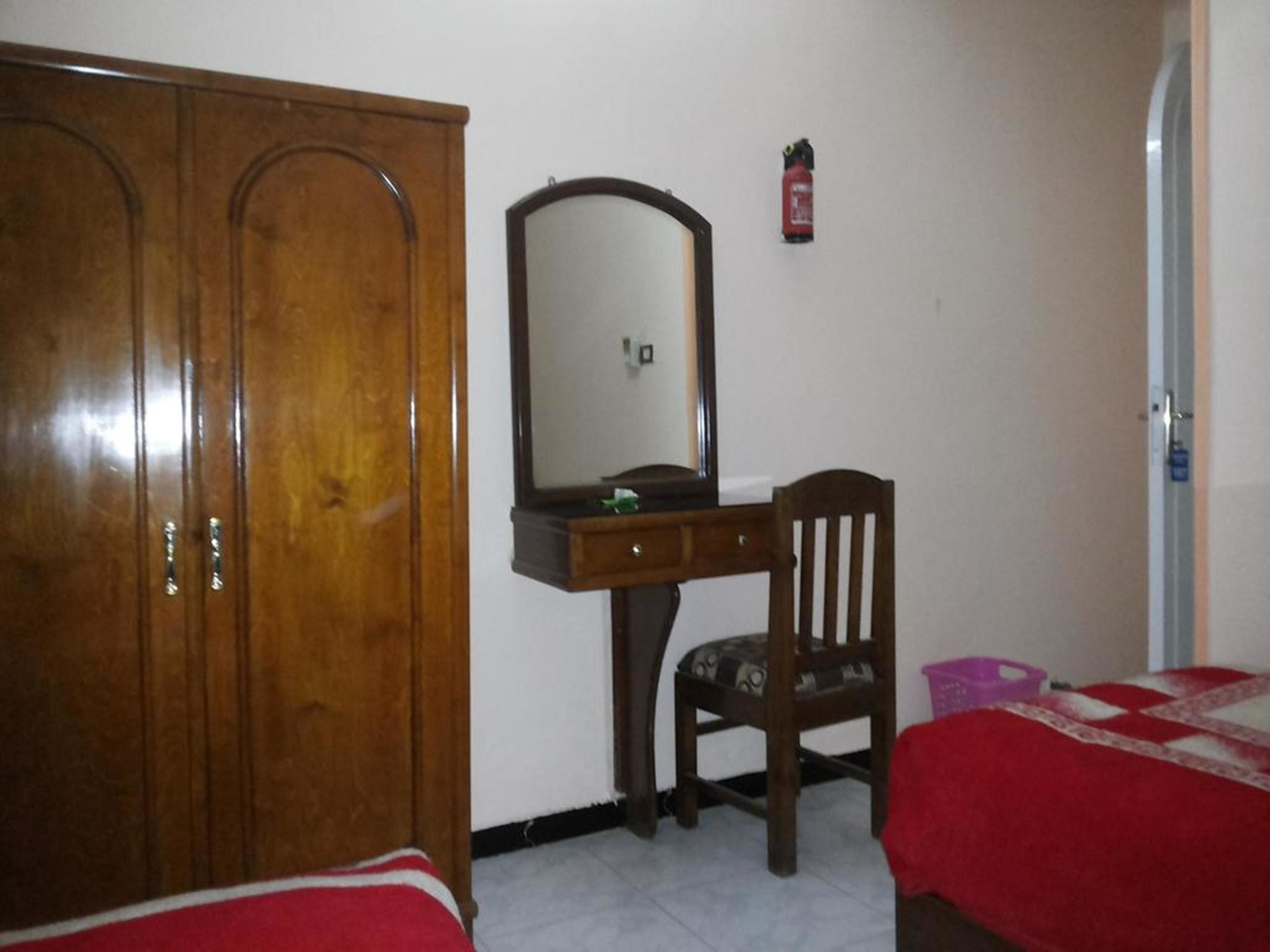 A bedroom with a wooden wardrobe, a desk with a mirror, a chair, and two beds with red bedding.