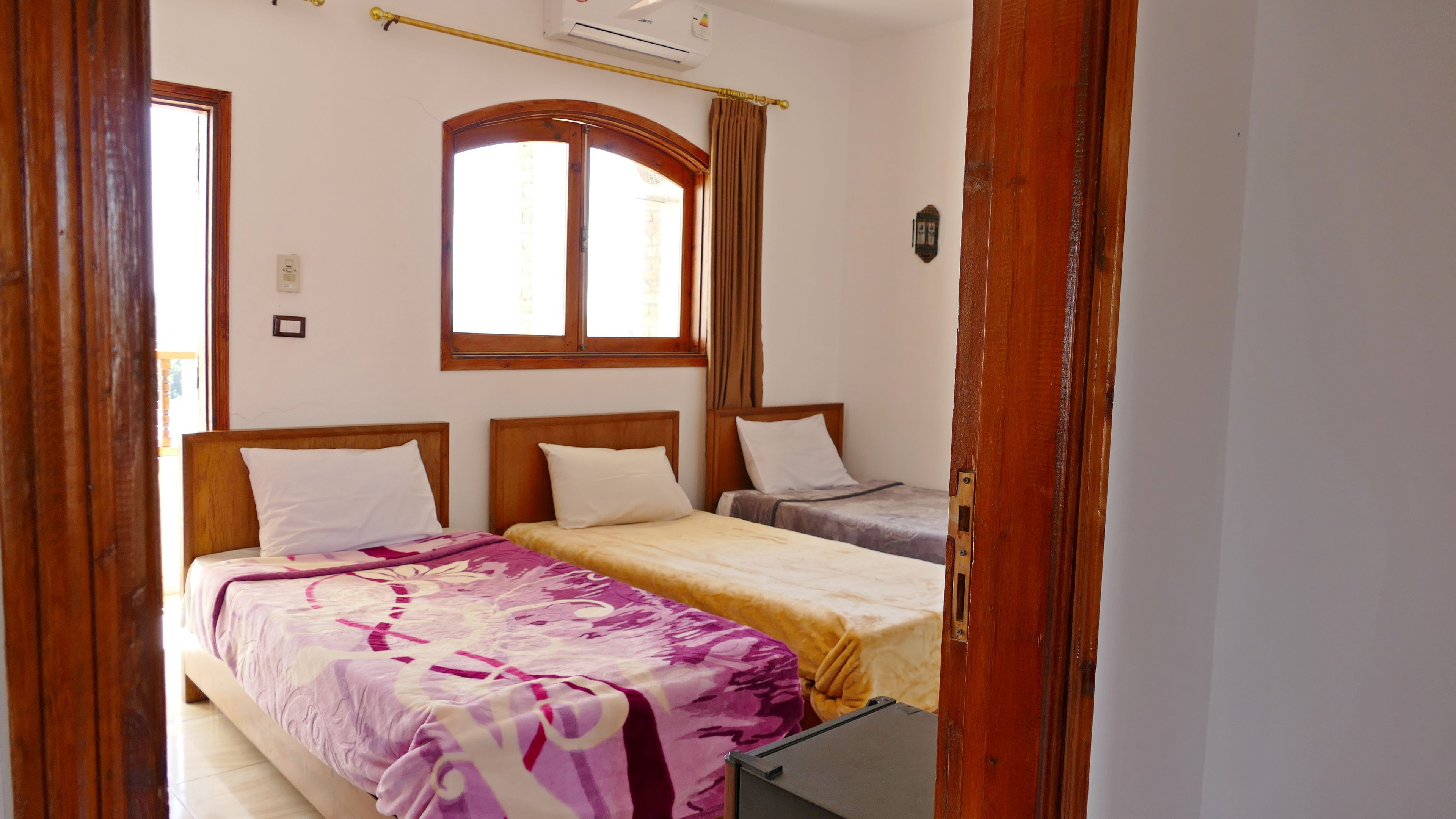 A room with two beds, wooden headboards, a window with curtains, and a wall-mounted air conditioning unit.