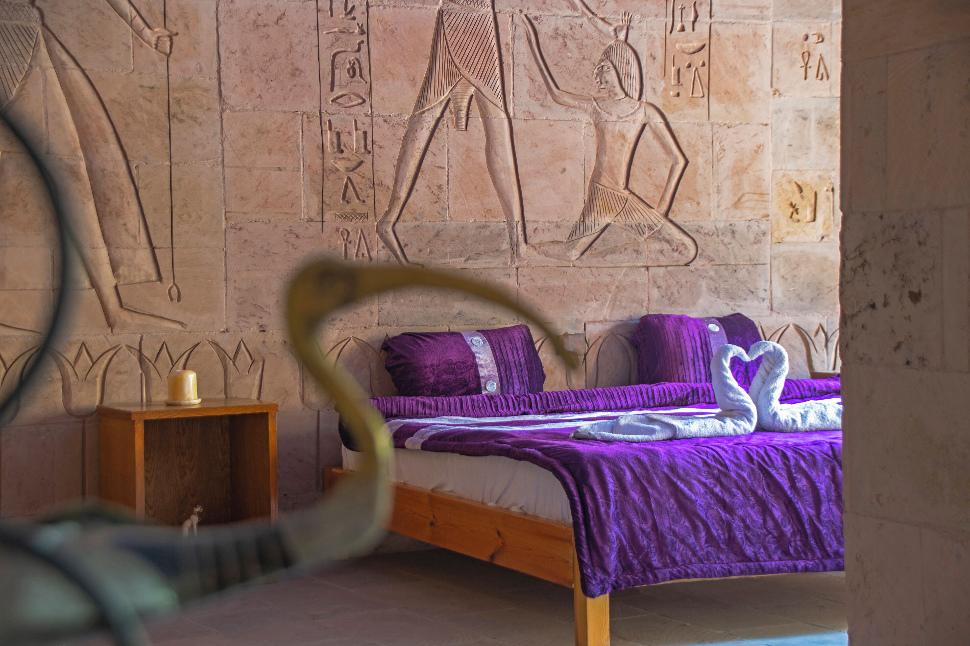 A room with a bed, a small wooden nightstand, and a wall with ancient Egyptian hieroglyphs.