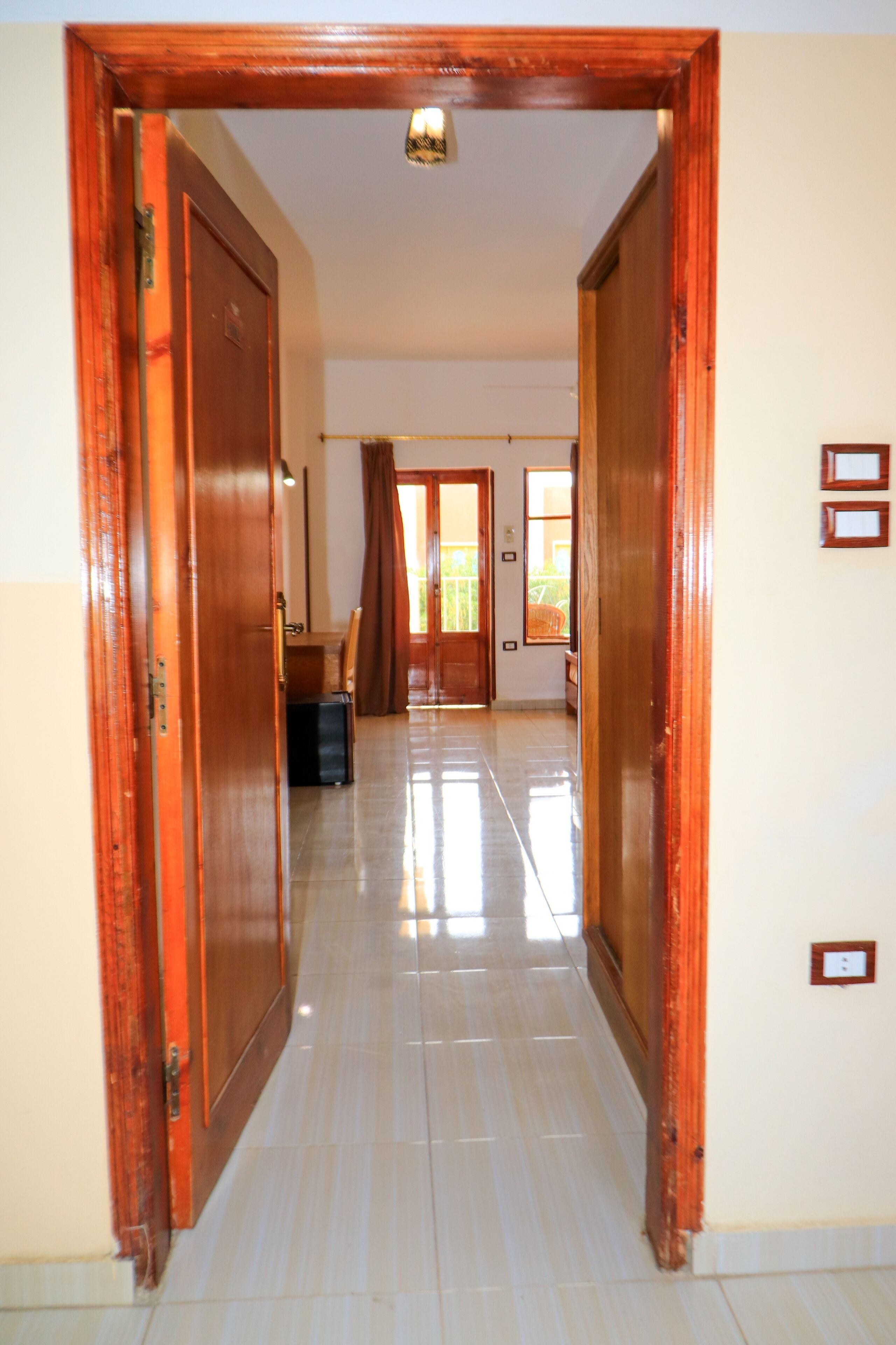 Un pasillo con puertas de madera, suelo de baldosas y vista a una habitación con cama y ventana.