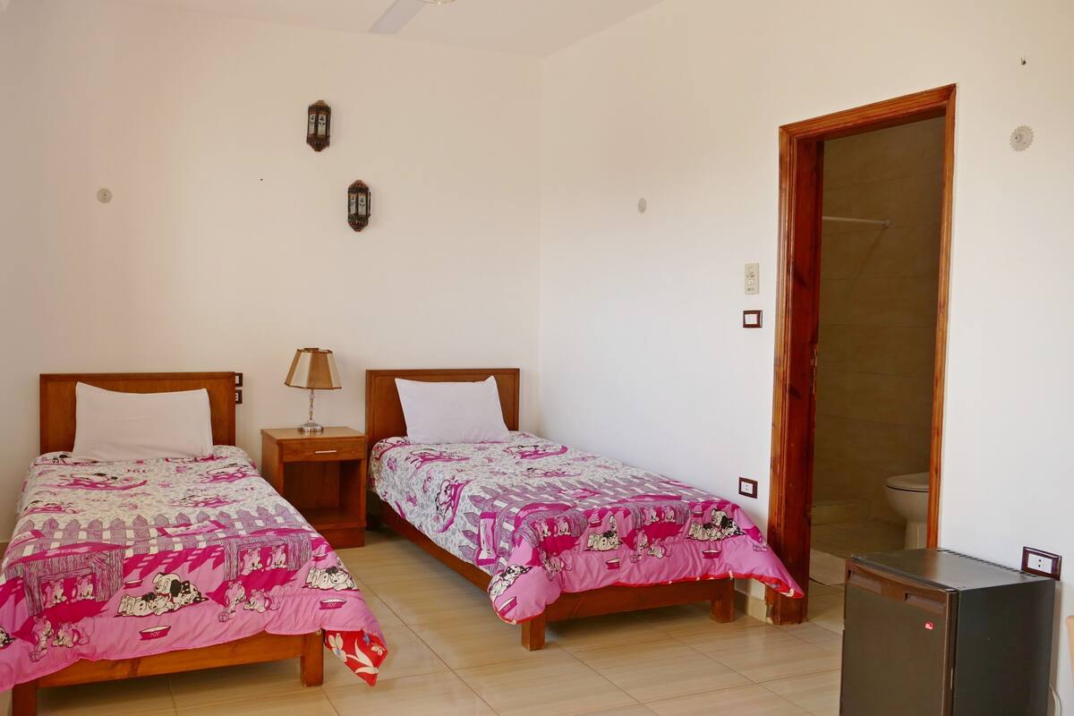 A room with two beds, a nightstand with a lamp, a wooden door, and a small cabinet.