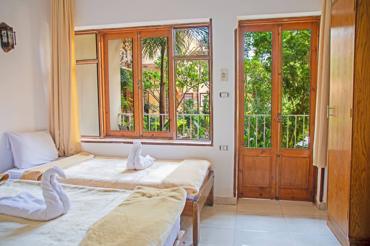A hotel room with two beds, a balcony with a view of greenery, and a wooden door.