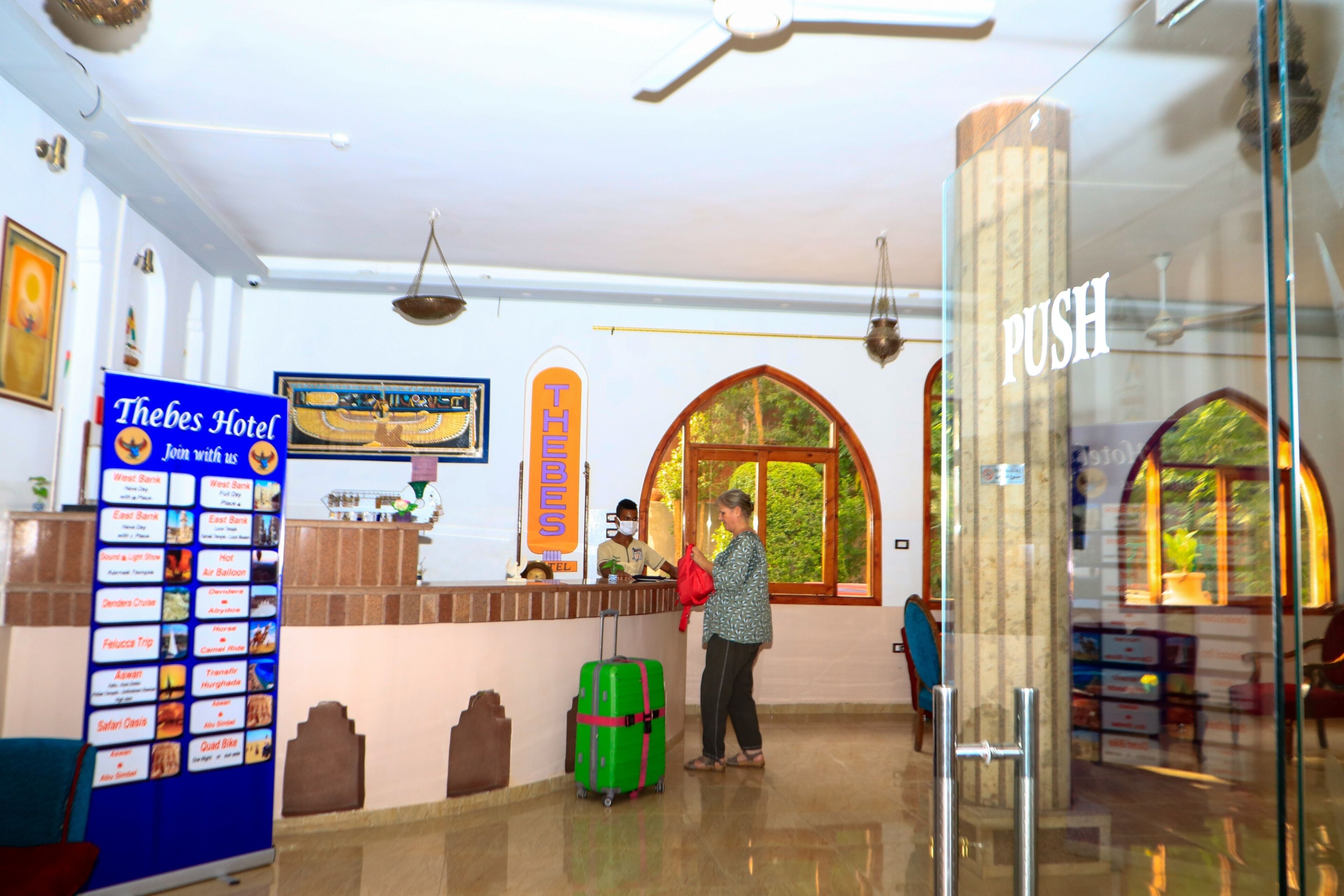 A hotel reception area with a reception desk, a 'PUSH' sign, a 'Thebes Hotel' signboard, and a large arched window.