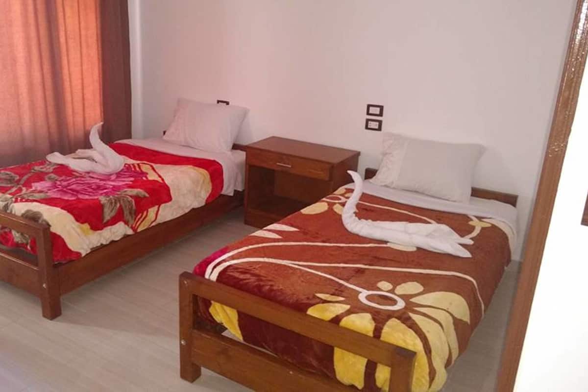 Two single beds with wooden frames, each with a red and yellow patterned bedspread and a white pillow. There is a wooden nightstand between the beds, and a window with curtains on the left.