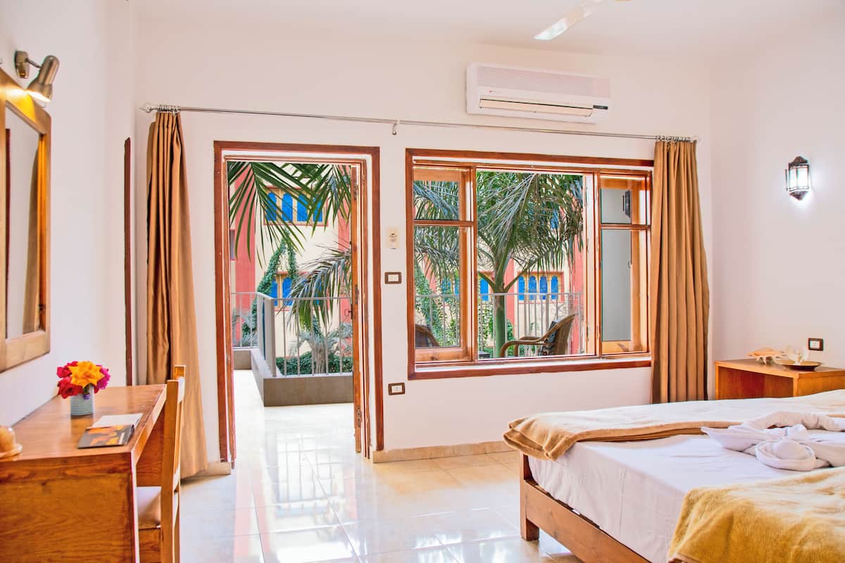A hotel room with a bed, a desk with a chair, a window with curtains, and a view of palm trees and buildings outside.