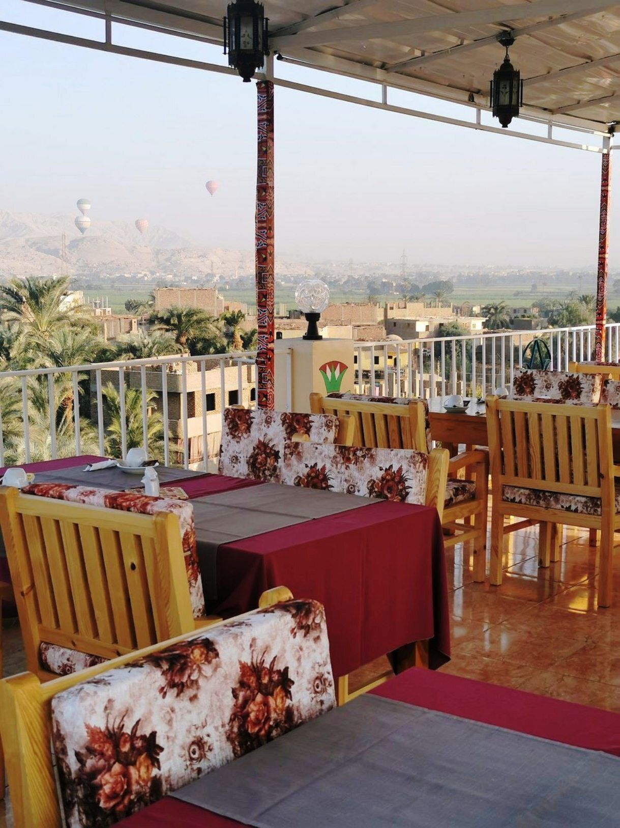 An outdoor dining area with tables and chairs, a view of the cityscape, and decorative elements like lanterns and patterned columns.