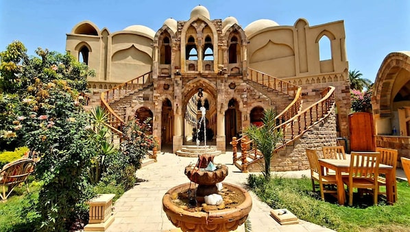 A historic building with a grand staircase, a central fountain, and lush greenery. - Embrace Hotel (Luxor)