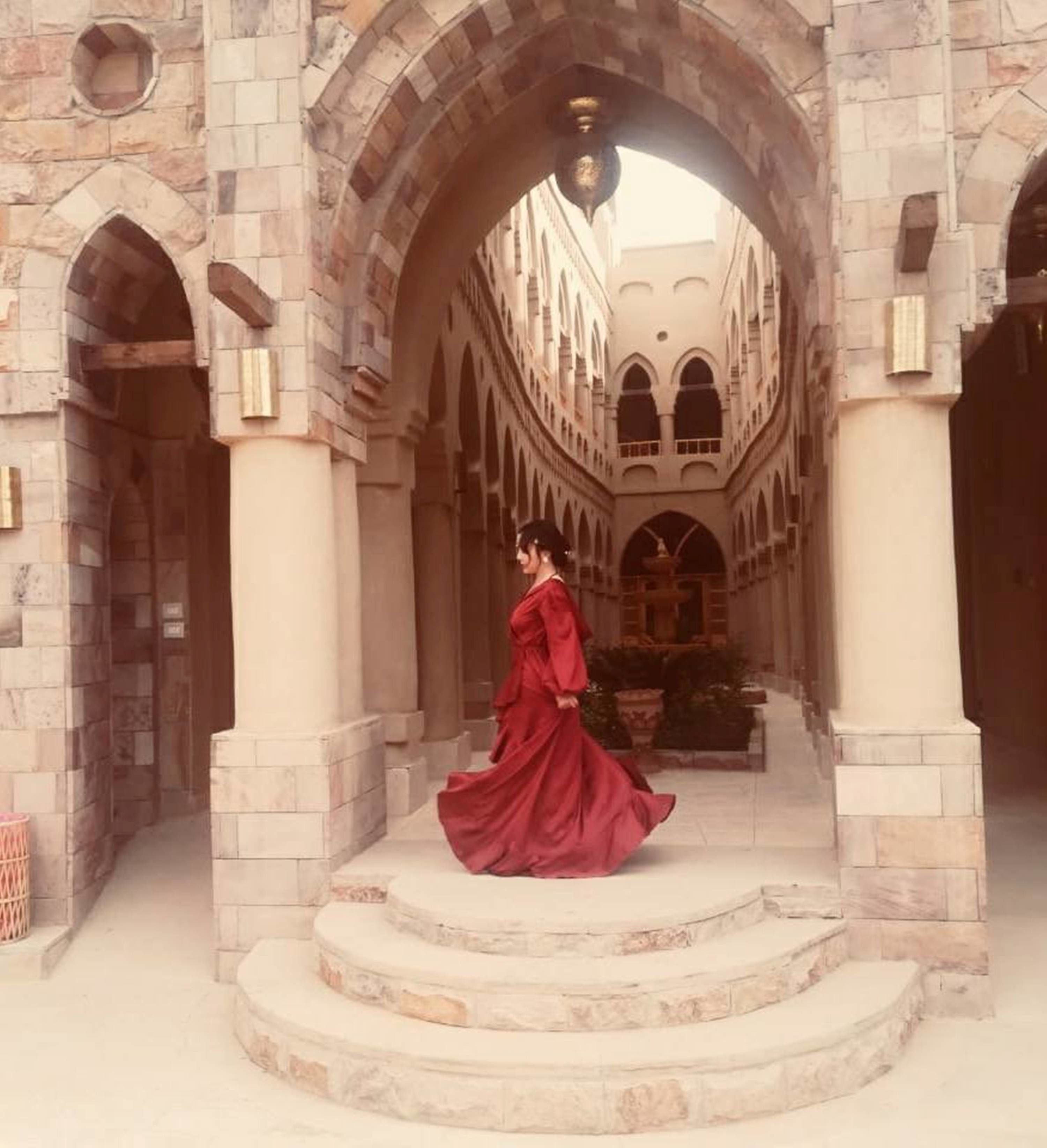 Una donna con un vestito rosso cammina in un lungo corridoio in pietra ad arco con gradini che conducono a un cortile.