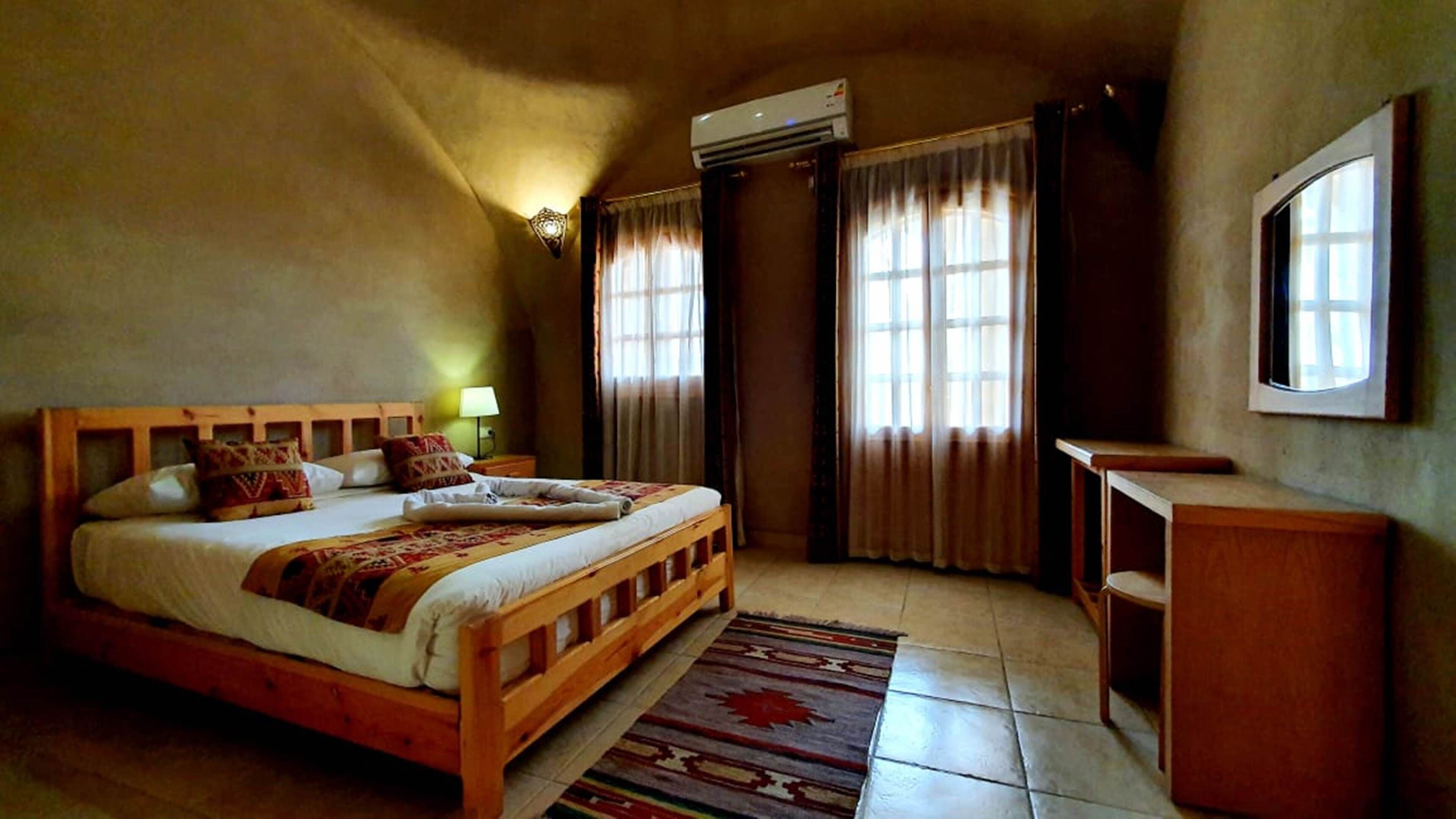 A bedroom with a wooden bed, a nightstand, a desk, a chair, a window with curtains, an air conditioning unit, and a wall-mounted TV.