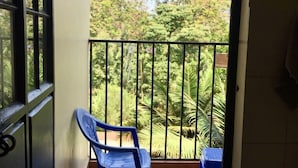 Un balcón con dos sillas de plástico azules y una mesita, con vista a un jardín exuberante con diversas árboles y plantas.