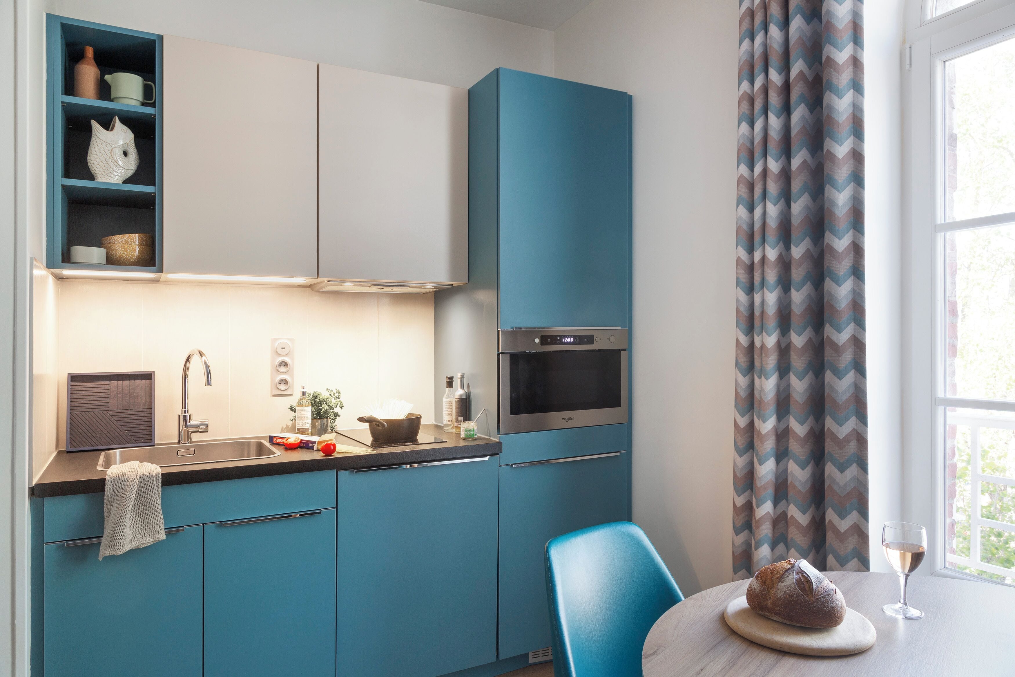 Eine moderne Küche mit taubenblauen Schränken, einem eingebauten Backofen und einem Fenster mit Zickzack-Vorhängen.
