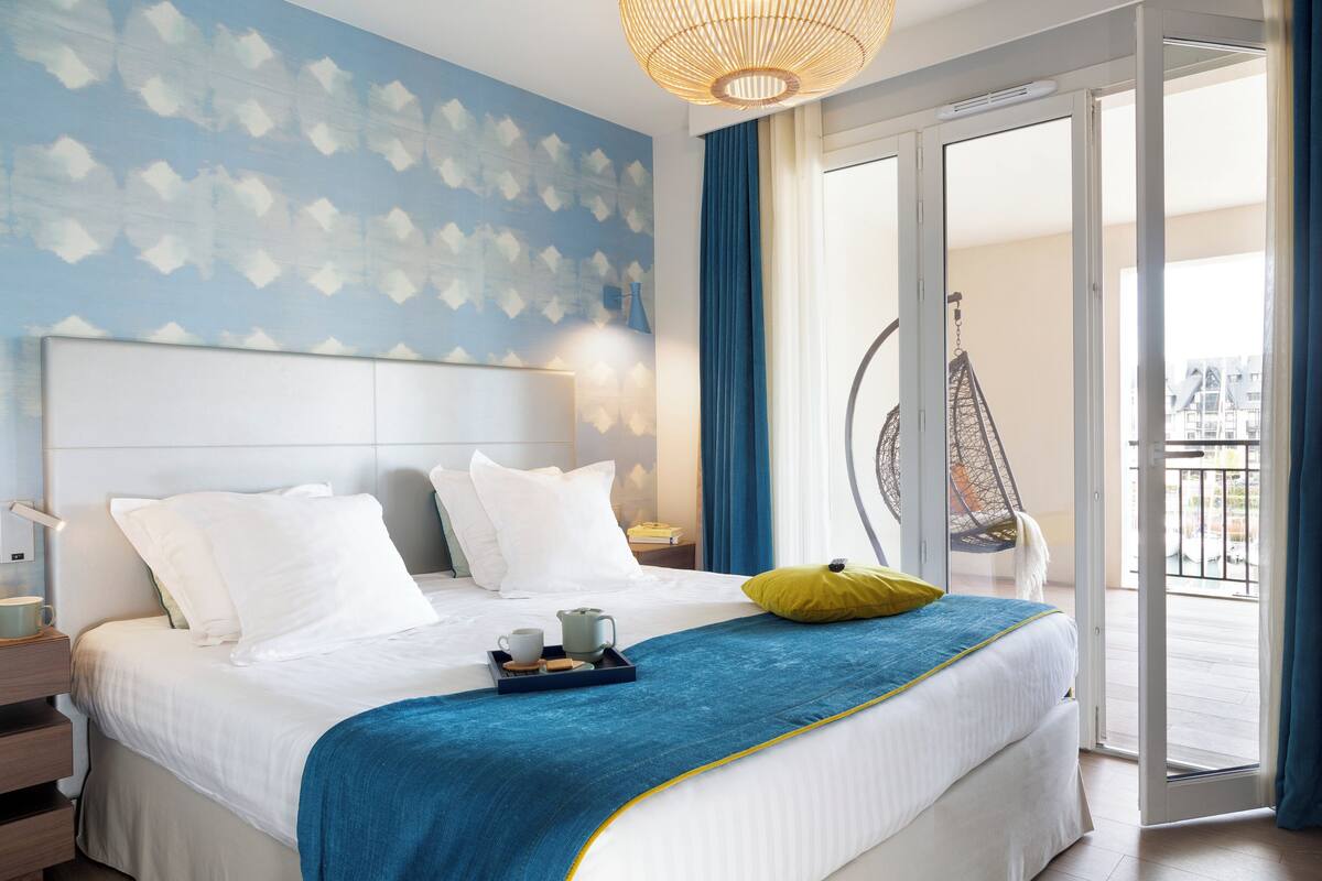 Quarto de hotel com cama, bandeja com xícara e biscoitos, cadeira suspensa e vista para o exterior.