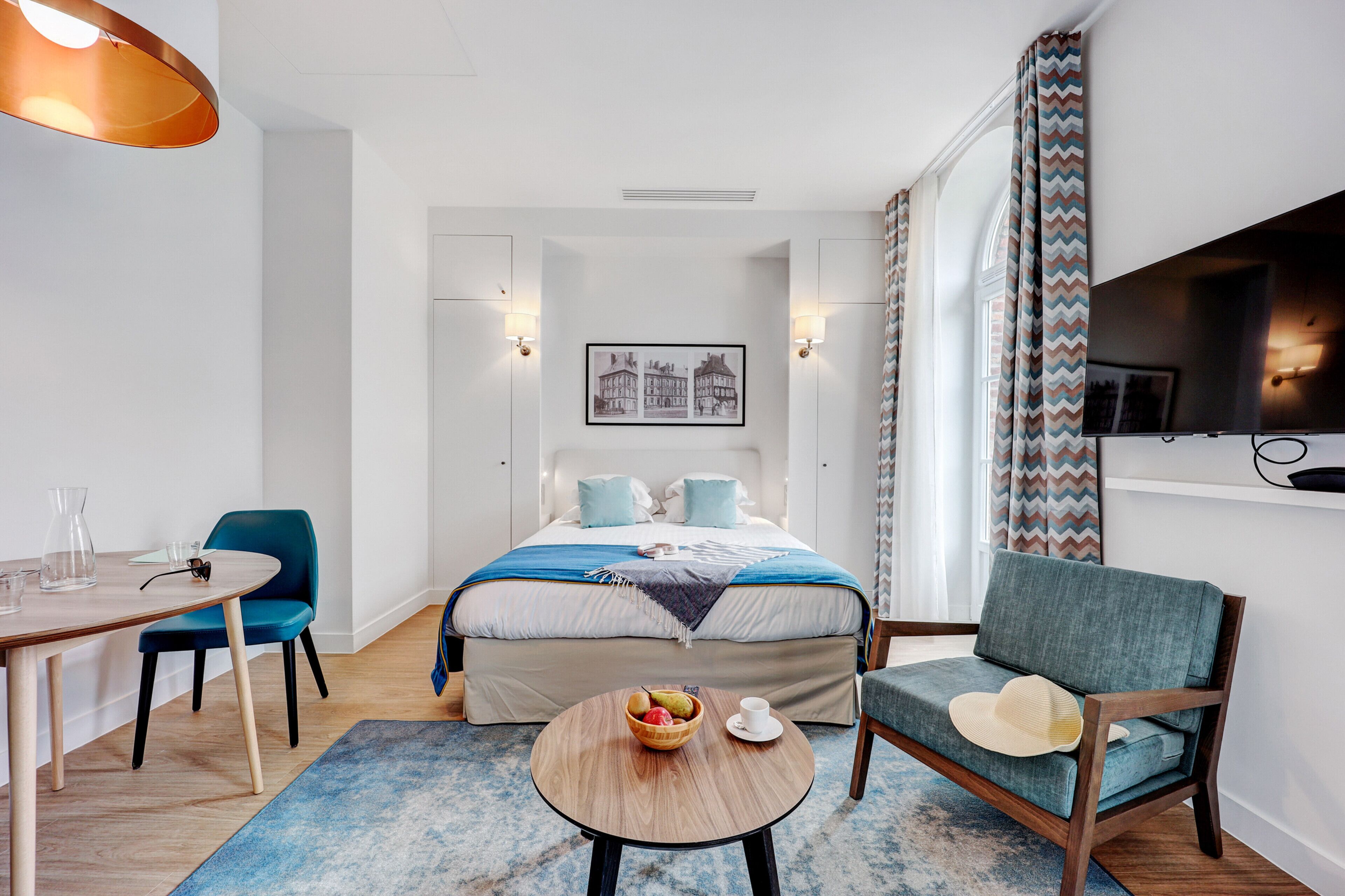 Moderni hotellihuone, jossa on sänky, työpöytä, tuoli, televisio ja ikkuna, jossa on verhot.