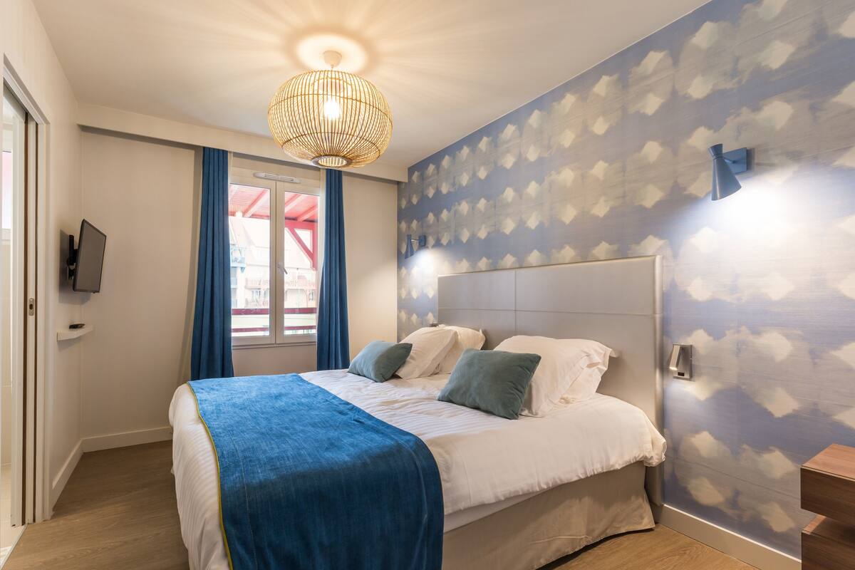 Quarto com cama, luminária pendente, parede com padrão de nuvens e janela com cortinas.