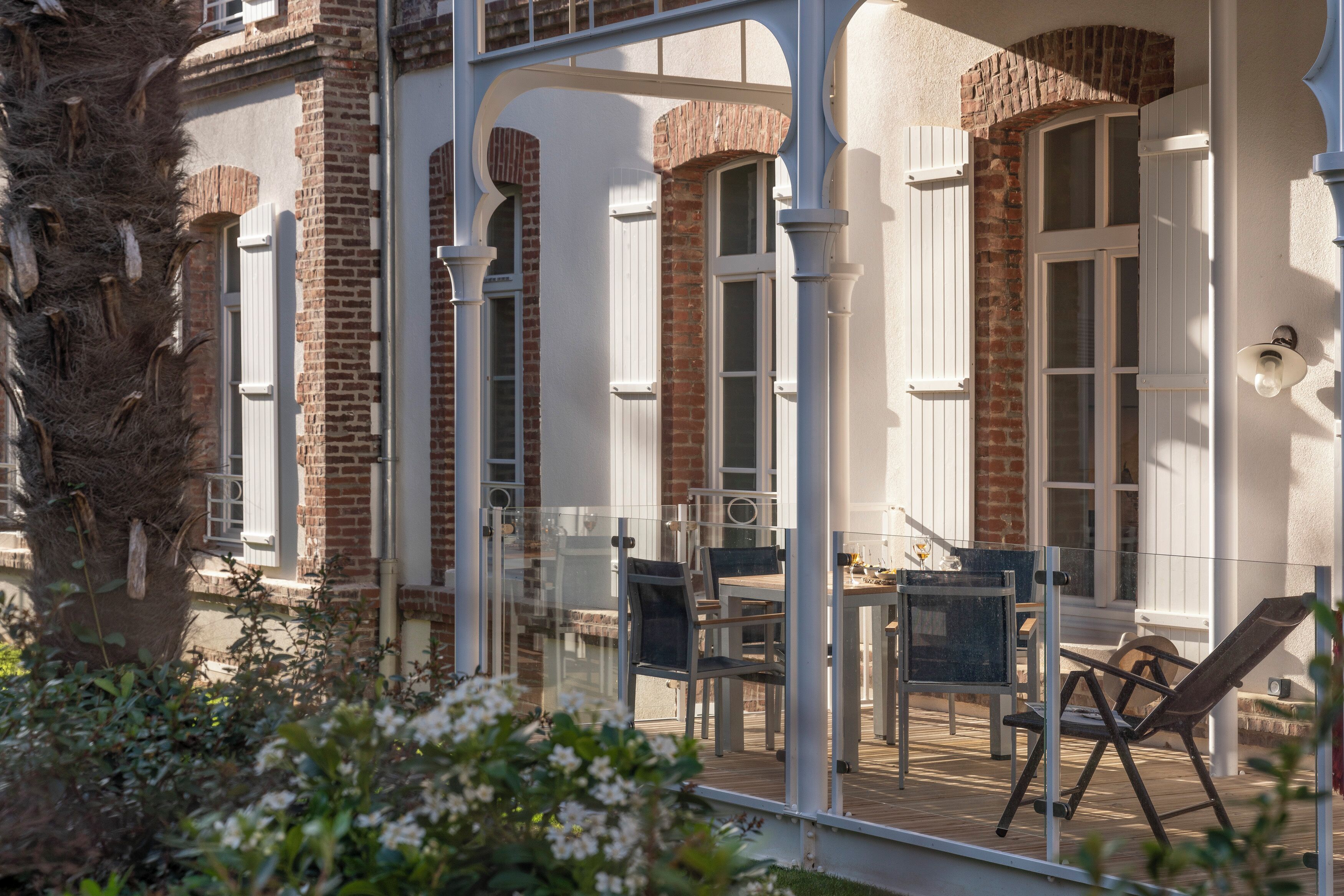 Een overdekt buitengebied met een tafel en stoelen, een houten terras en een bakstenen gebouw met witte luiken.