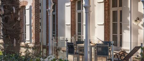 A covered outdoor area with a table and chairs, a wooden deck, and a brick building with white shutters.