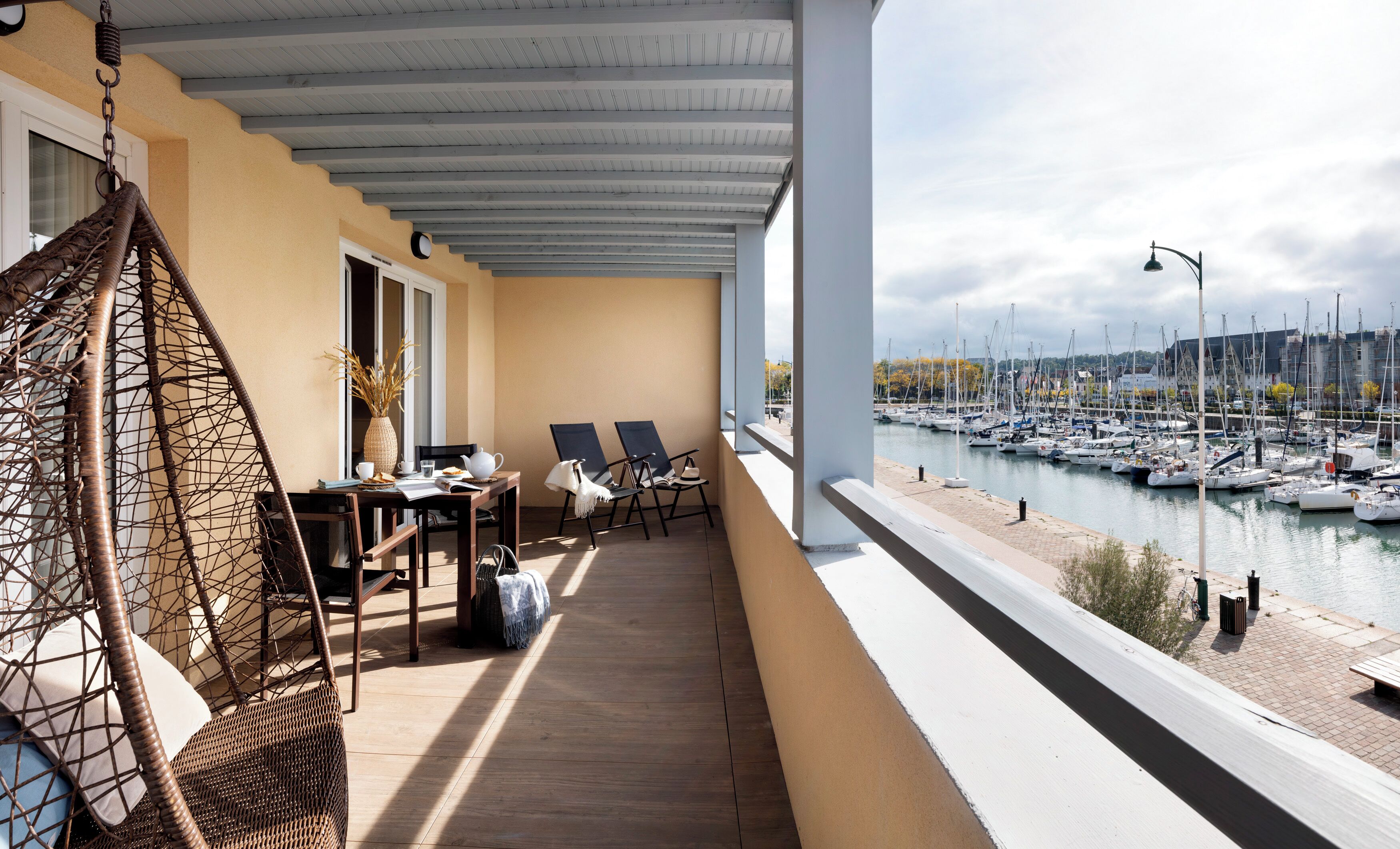 Ein Balkon mit Korbstühlen, einem Tisch mit einer Vase und Blick auf eine Marina mit Booten.