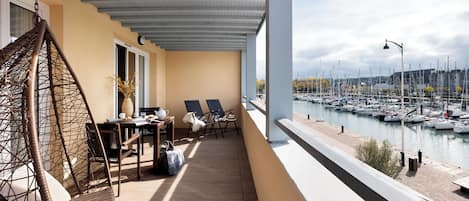 Een balkon met rieten stoelen, een tafel met een vaas, en uitzicht op een jachthaven met boten.