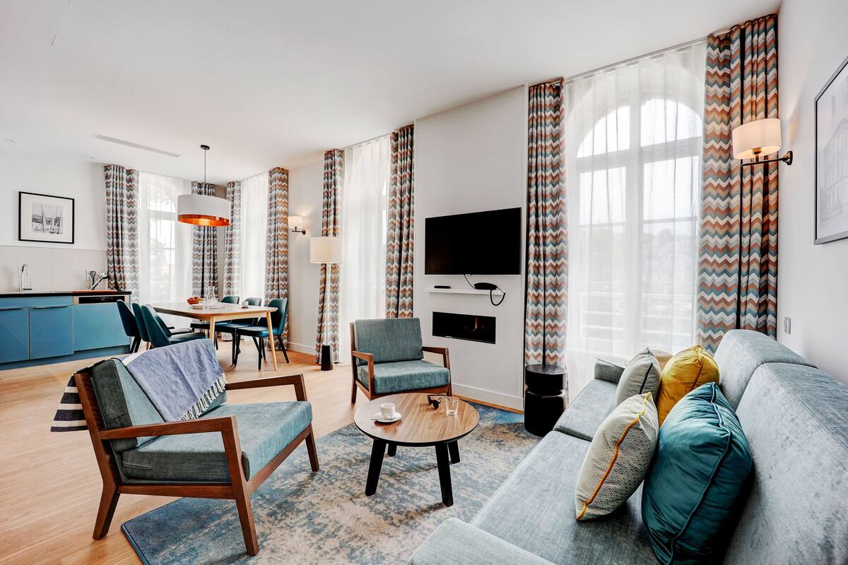 Uma sala moderna com um sofá, poltronas, uma mesa de centro e uma TV. Há uma área de jantar com uma mesa e cadeiras, e uma cozinha com armários azuis.