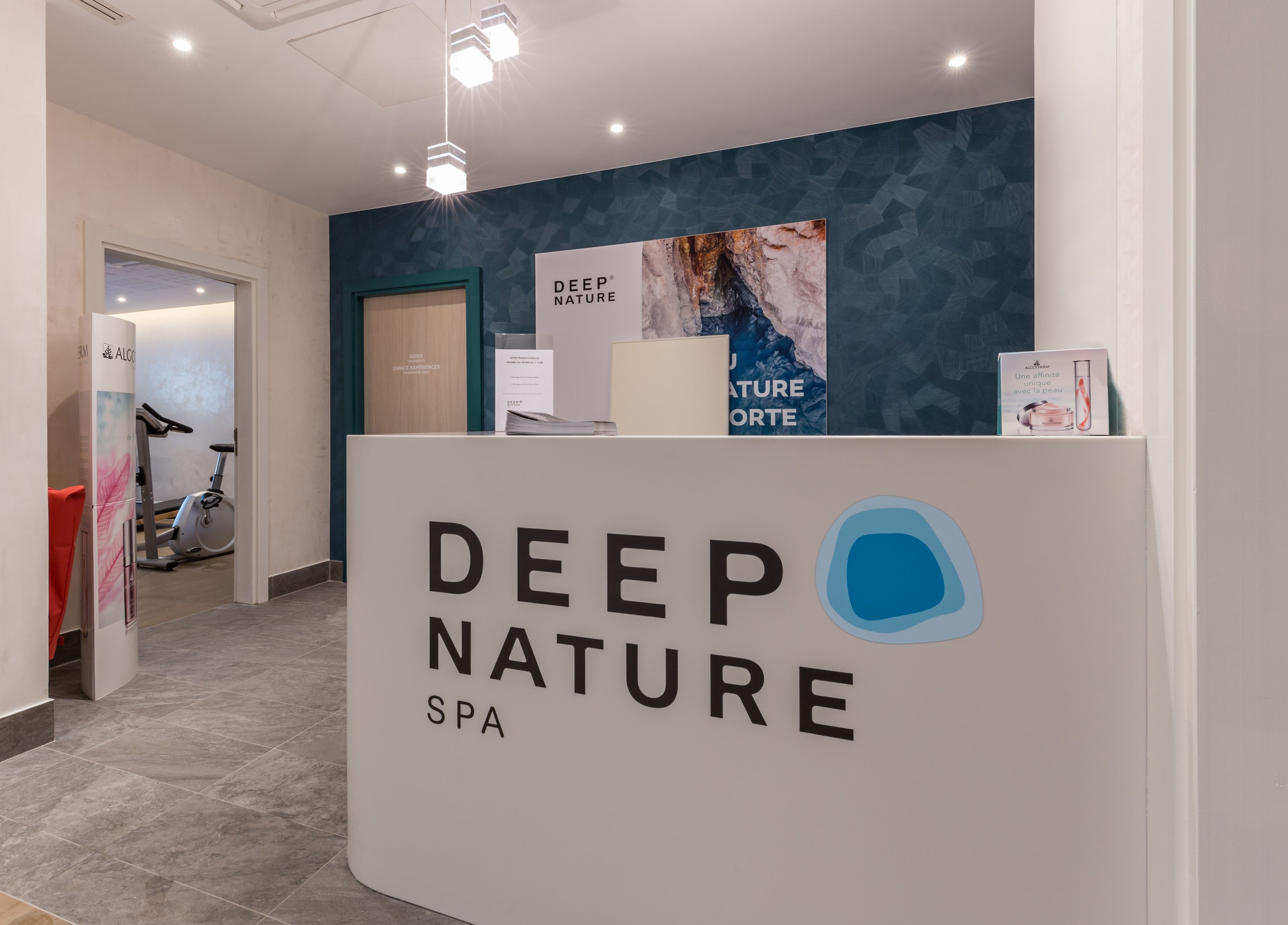 A reception area with a sign for Deep Nature Spa, a wall with abstract art, and a glimpse of a gym in the background.