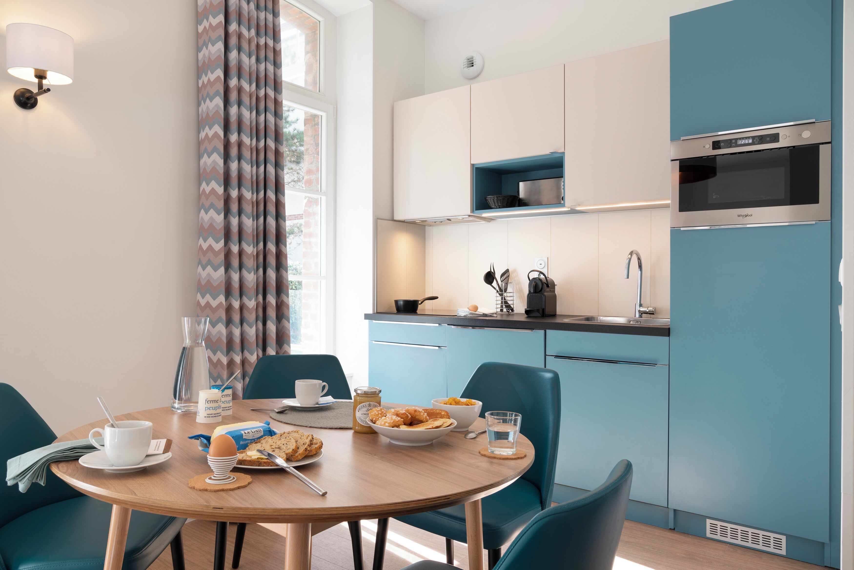 A modern kitchen with a microwave, sink, and blue cabinets. There is a dining table with breakfast items and chairs.