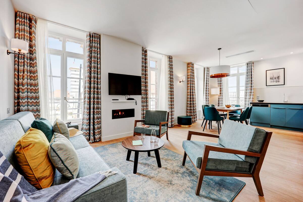 Uma sala de estar moderna com sofá, poltronas, mesa de centro e televisão. Há uma área de cozinha com um armário azul e uma mesa de jantar com cadeiras.