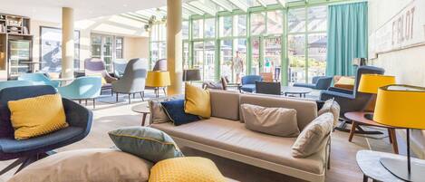 A spacious lobby with a modern design, featuring a central seating area with sofas and armchairs, a large glass wall providing a view of the outside, and a well-lit environment with natural light.
